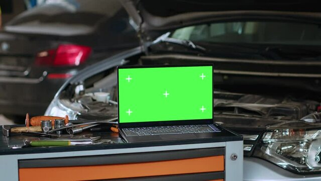 Close up panning shot of green screen laptop in auto repair shop, sitting on work bench next to professional mechanic tools in front of car with hood up waiting to be repaired