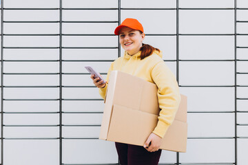 Young woman in red hat picks up parcel from automatic post office machine, Courier standing with...