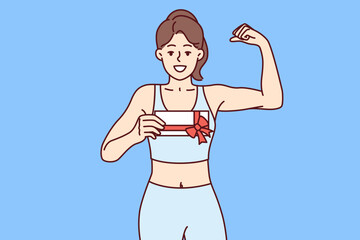 Woman holds gift certificate for fitness club and shows biceps, urging you to do sport. Smiling girl demonstrates fitness voucher with red ribbon presented for birthday and motivating to visit gym