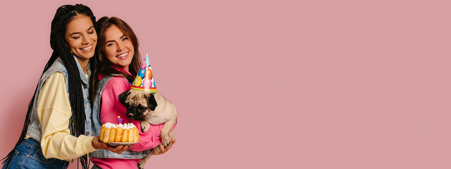 Two joyful young women bonding while carrying cute pug dog and birthday cake against pink background