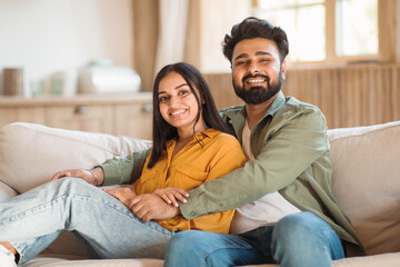 Happy indian lovers enjoying time together at home, sitting on couch, embracing and smiling at camera, free space