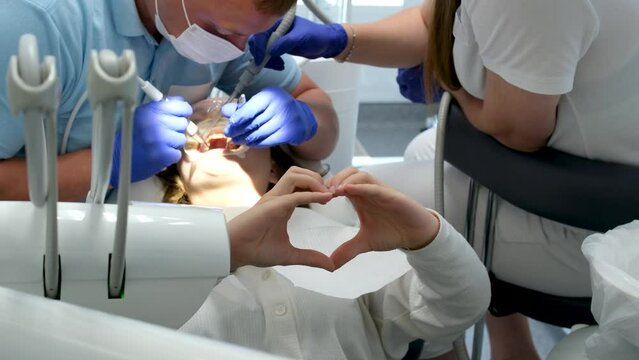 young girl showing heart with hands Sitting in dental office brushing teeth whitening dental procedures doctor and nurse saliva suction and other latest technologies in blue gloves and masks.self care