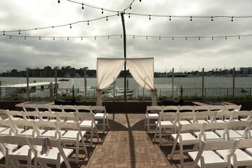 an outdoor wedding setup has white chairs by the water with lights strung above it