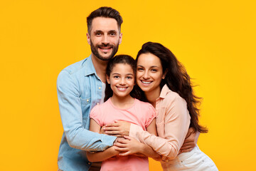 Happy european parents hugging their pretty daughter and smiling to camera on yellow background,...