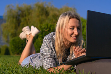 Portrait of a young smiling blonde woman lying on her stomach on the grass in the park with a laptop on a sunny day. Woman communicates, works on a laptop in a city park