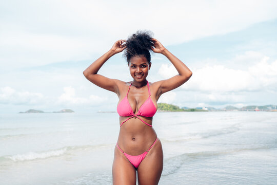 Beautiful African american woman in pink bikini on tropical beach. Portrait of dark skinned woman smiling at sea. Brunette tanned girl in swimwear enjoying and walking on beach.