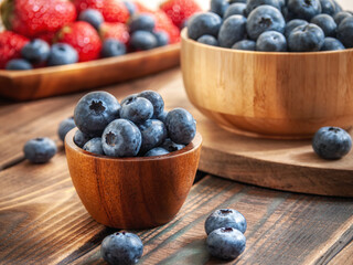Blueberries close-up in a wooden cup on a dark brown wooden background in warm rays of light. Blueberries and strawberries in the background. Selective focus. Side view.