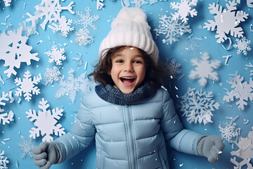 Top view of a child in a winter down jacket and a knitted hat lying on a blue background with snowflakes.
