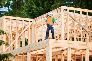 Carpenter building wooden frame two-story house. Bearded man wearing protective goggles carrying...