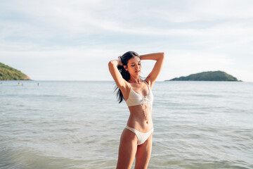 Beautiful caucasian woman in white bikini on tropical beach. Portrait of dark skinned woman smiling at sea. Brunette tanned girl in swimwear enjoying and walking on beach.