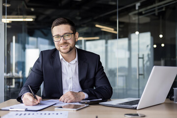 Portrait of a young businessman smiling at the camera, sitting at a desk in the office and working with documents and an invoice - Powered by Adobe