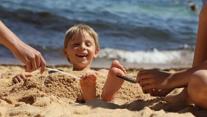 Child, tickling sibling on the beach on the feet with feather, kid cover in sand, smiling, laughing, enjoying some fun - Powered by Adobe