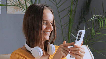 Cheerful smiling brown haired woman holding mobile phone chatting online with friends posing with headphones over neck, smiling reading good news.