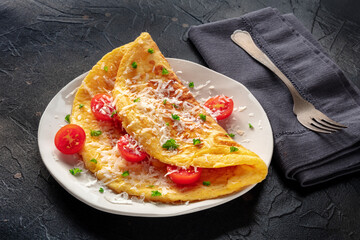 An omelet with tomato and parsley, eggs for breakfast, a healthy vegetarian dish with cheese, on a black slate background, with a fork - obrazy, fototapety, plakaty