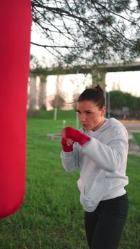 Kickboxing woman in hoodie hitting punching bag while boxing workout in park. Athletic female boxer hands wrapped in red boxing tapes training outdoor. Female fitness motivation. Vertical video