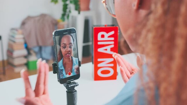 Young professional attractive African American woman records video on mobile phone camera with tips for fans or answers questions from subscribers live sits at table with on air sign in office.