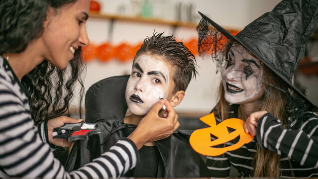 Close-up portrait of three people preparing for Halloween. An Oriental mother paints the face of her little son and makes him up like a vampire, her daughter, a girl, sits nearby in a witch costume.