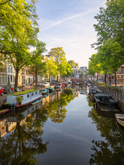 This photo showcases a peaceful Amsterdam canal scene. It includes an arched bridge, trees, boats...