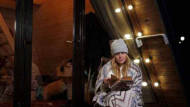 A woman is sitting in an armchair wrapped in a blanket and reading a book on a cold autumn evening near a small cozy house. A rustic wooden house with a triangular roof descending to the ground.