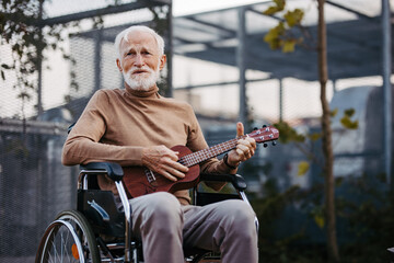 Senior man sitting in a wheelchair outdoors, playing the ukulele.