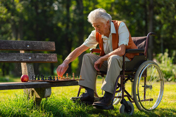 A senior man in a wheelchair with chess on bench is alone in a city park, feeling lonely. Elderly...