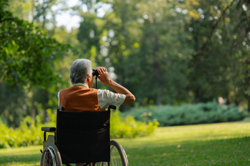 Senior man in wheechair spending free time outdoors in nature, watching forest animals through...