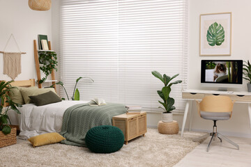 Comfortable bed, desk with computer and potted houseplants in stylish bedroom. Interior design