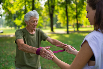The senior man exercising with physiotherapist in the park, using resistance band. Elderly healthcare.
