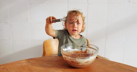 Toddler stirs chocolate pancake batter in large bowl, proudly raising whisk high, showcasing his culinary prowess. Heartwarming moment of learning and achievement in kitchen, captured in slow motion