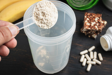 Male hand puts into a shaker scoop portion of whey, soy protein powder. White capsules of amino acids, vitamins, protein chocolate bar and bananas on a dark wooden board, bodybuilding food supplements