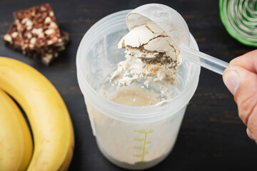 Male hand puts into a shaker scoop portion of whey, soy protein powder. Protein chocolate bar and bananas on a dark wooden board, bodybuilding food supplements
