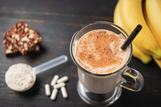 Glass of protein milkshake drink with straw and scoop of whey protein powder, white capsules of amino acids, vitamins, protein bar and bananas on a dark wooden board, bodybuilding food supplements