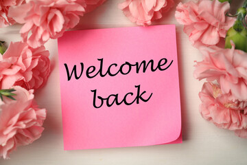Paper note with phrase Welcome Back and flowers on light table, flat lay