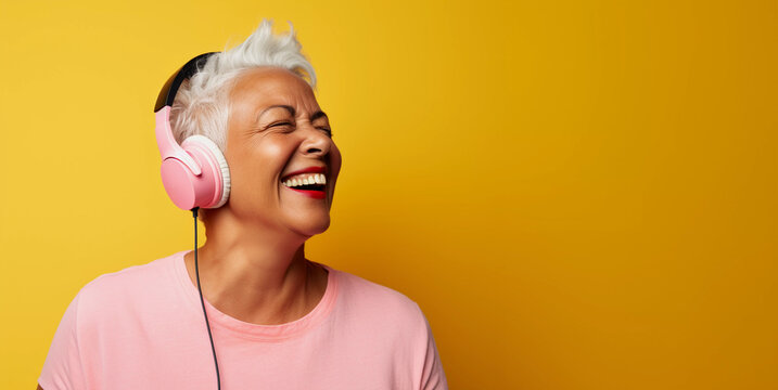 Studio portrait of middle aged black woman listening on headphones and laughing, colorful pink outfit and yellow background