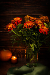 Bunch or bouquet of Tagetes flowers of orange colour with green leaves and stem in glass vase on textile napkin on the table with glowing candle against wooden wall is a symbol of Day of the Dead