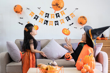 A playful young mother sits on a comfortable sofa in the living room decorated with pumpkins and her cute little daughter tells a spooky Halloween story. Funny mother and girl having fun at home