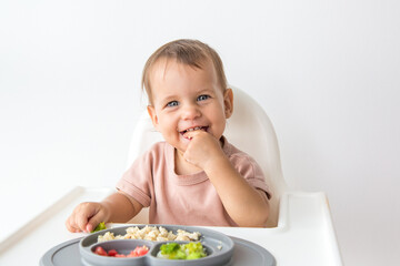 cute 1 year old baby sitting in a high chair and eating complementary foods, smiling and looking at...