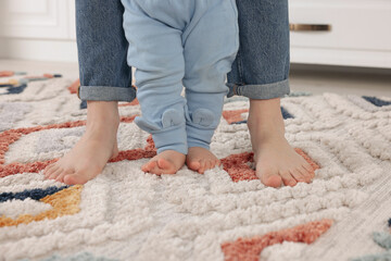 Mother supporting her baby son while he learning to walk on carpet indoors, closeup