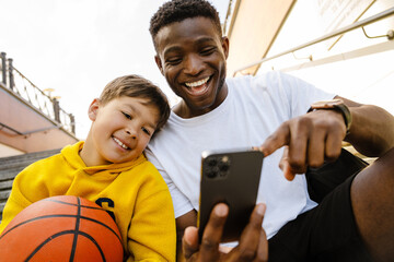 African man and his son using mobile phone while sitting outdoors after basketball training