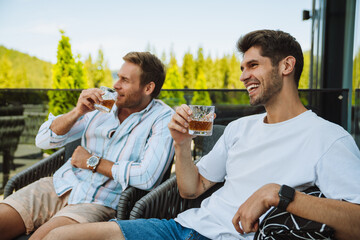 Two male friends drinking whiskey while relaxing on terrace in mountains