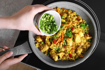 Woman adding cut green onion to rice with meat and vegetables in frying pan, top view