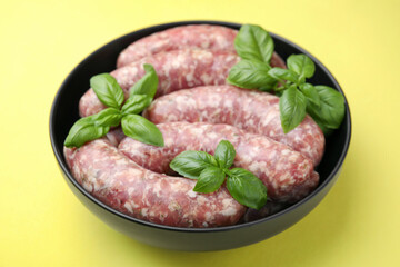 Raw homemade sausages and basil leaves in bowl on yellow background
