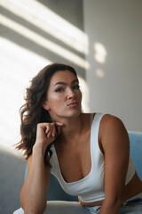 Beautiful brunette woman in white top and blue jeans sitting on blue armchair. Girl smile, happy. Portrait of young pretty woman. Smile and freshness. White background. Light from window, shadows