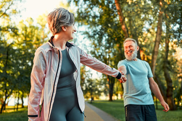 An older beautiful woman holds a man's hand and pulls him to sport training, and he stops and laughs. Lifestyle of the elderly. Activities in the fresh air.A healthy body is a healthy mind.