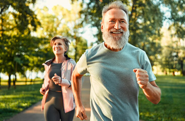 Cardio training. Close up of energetic fit sportsman spending morning in park and jogging together with wife. Active pensioners taking care of health and doing regular physical activities on nature. - obrazy, fototapety, plakaty