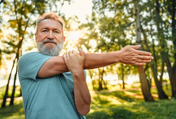 Sportsman working out on nature. Portrait of charismatic athletic man of mature age doing arms exercises for warming up outdoors. Healthy man dressed in blue shirt confidently looking at camera.