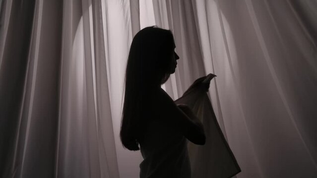 Medium slow motion video of a young woman's silhouette just out of the shower, taking a towel off her body, turning her back on a curtain background in a muffled light.