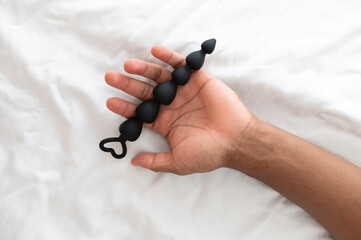 Black sex toy - anal beads in a hand of a young African man lying on the bed - obrazy, fototapety, plakaty
