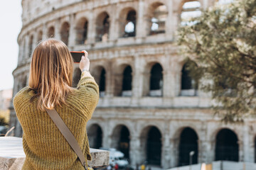 Fototapeta na wymiar Portrait of a woman on background of Coliseum in Rome, Italy. Beautiful female in dress using smartphone outdoors. Traveler. Female tourist taking photo on mobile phone of old ruins