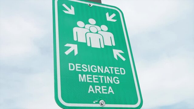 designated meeting area vertical rectangle green and white sign on post with sky behind, illustration of people with four arrows pointing at them